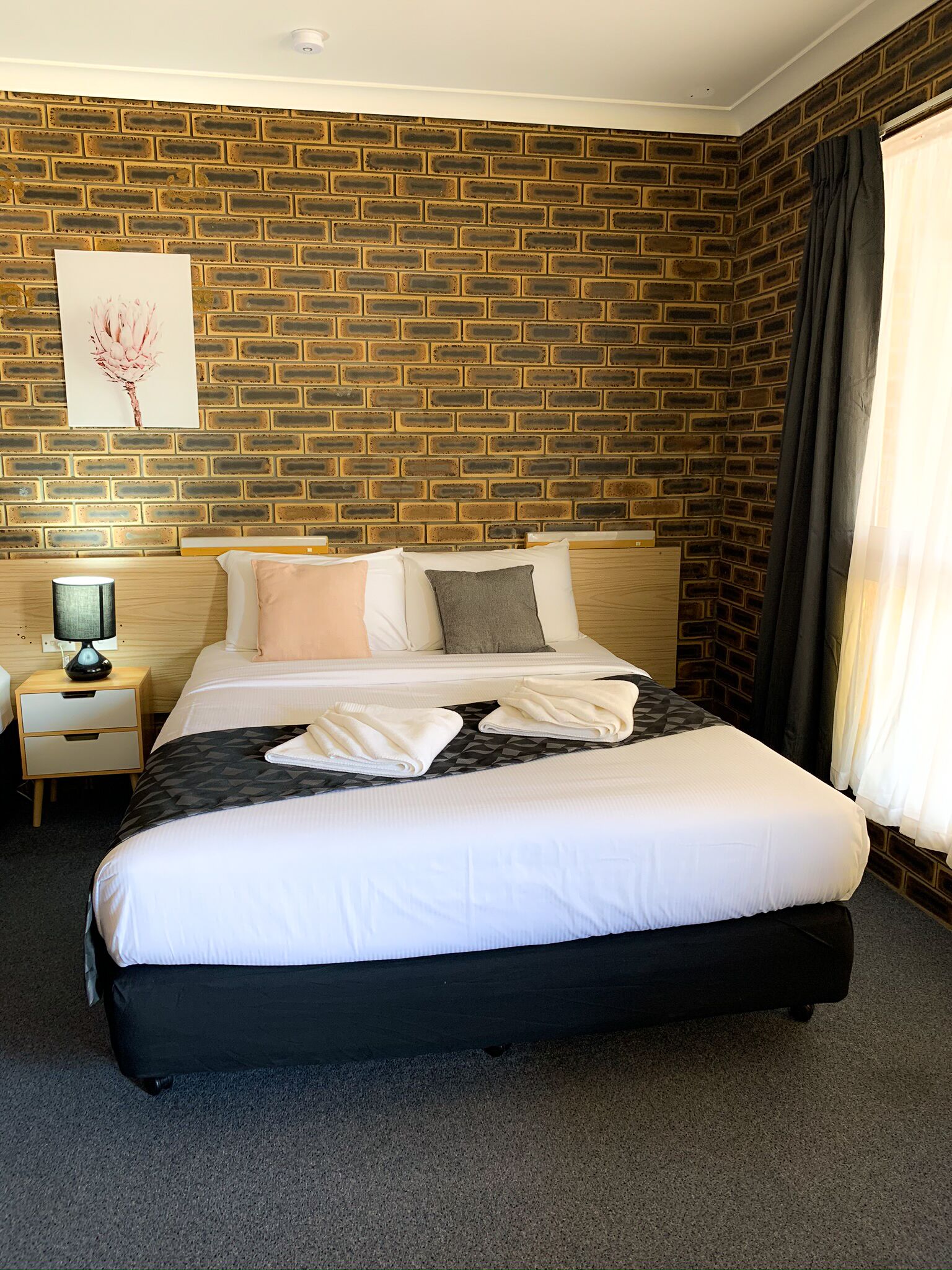 Una habitación de hotel con pared de ladrillo, una cama bien tendida con ropa de cama blanca y gris, una mesita de noche de madera con una lámpara, y un cuadro en la pared.