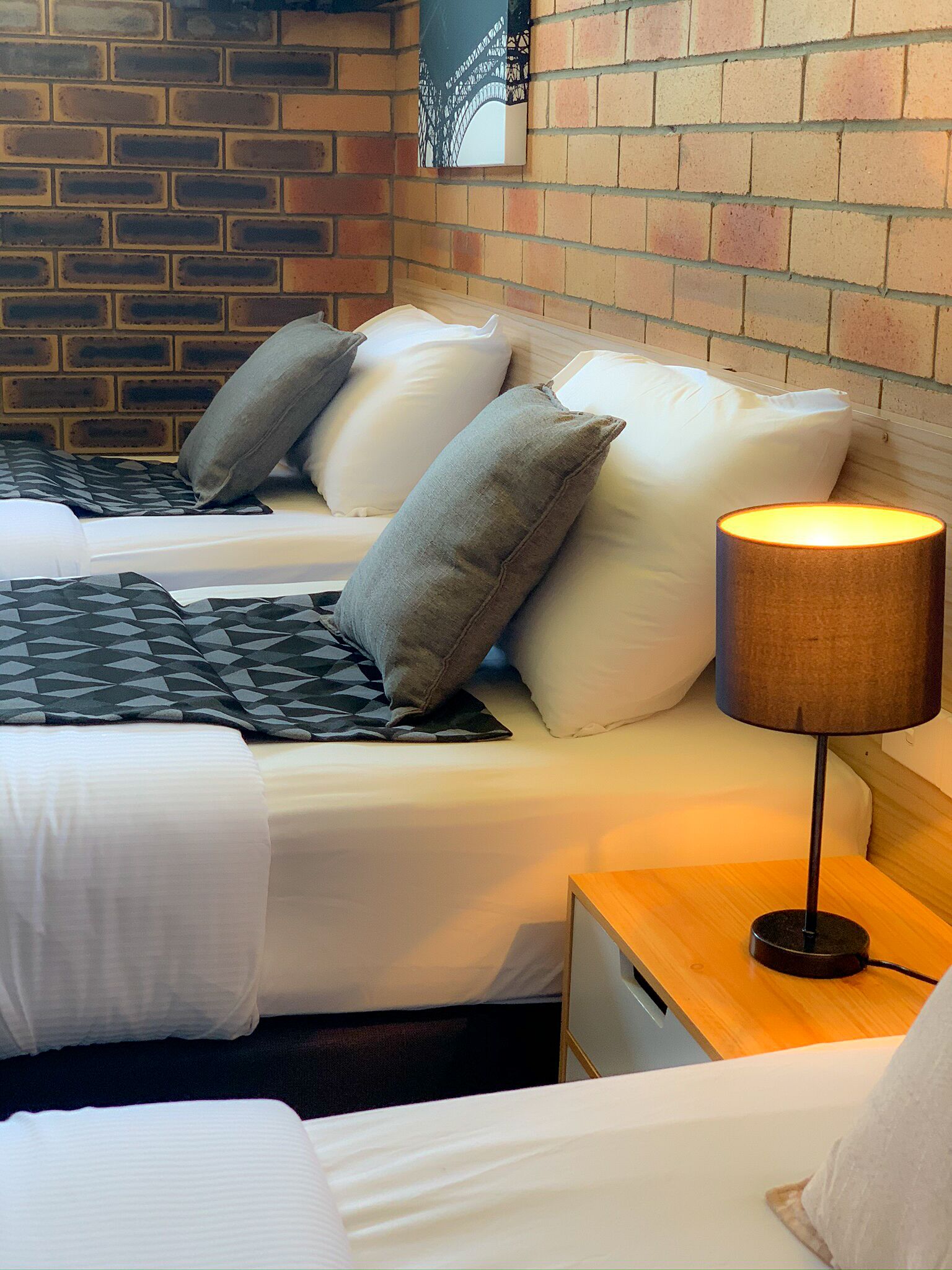 A double bed with white bedding and grey pillows, a lamp on a wooden nightstand, and a brick wall with a decorative item.