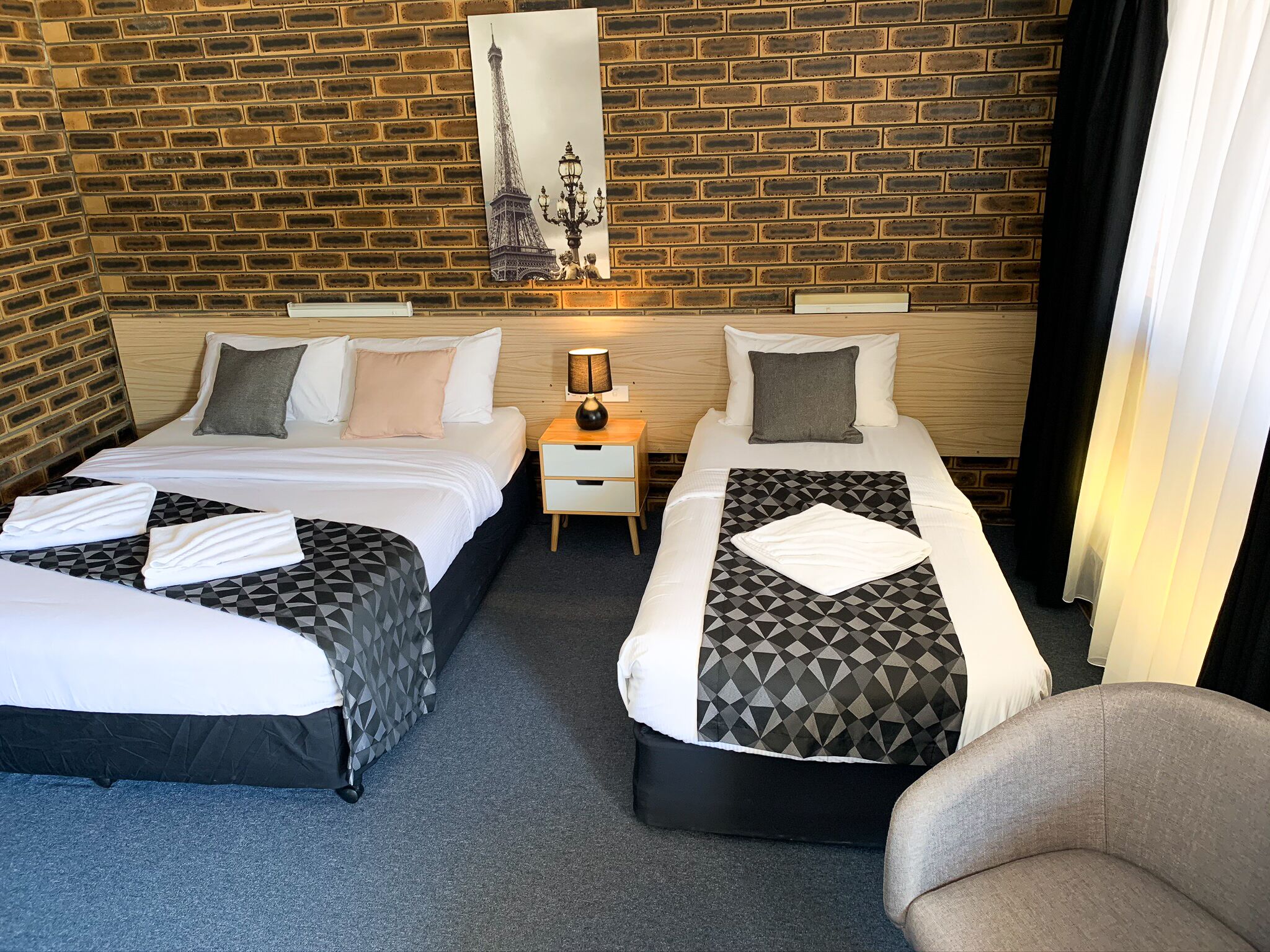 A hotel room with two beds, a nightstand, a lamp, and a brick wall.