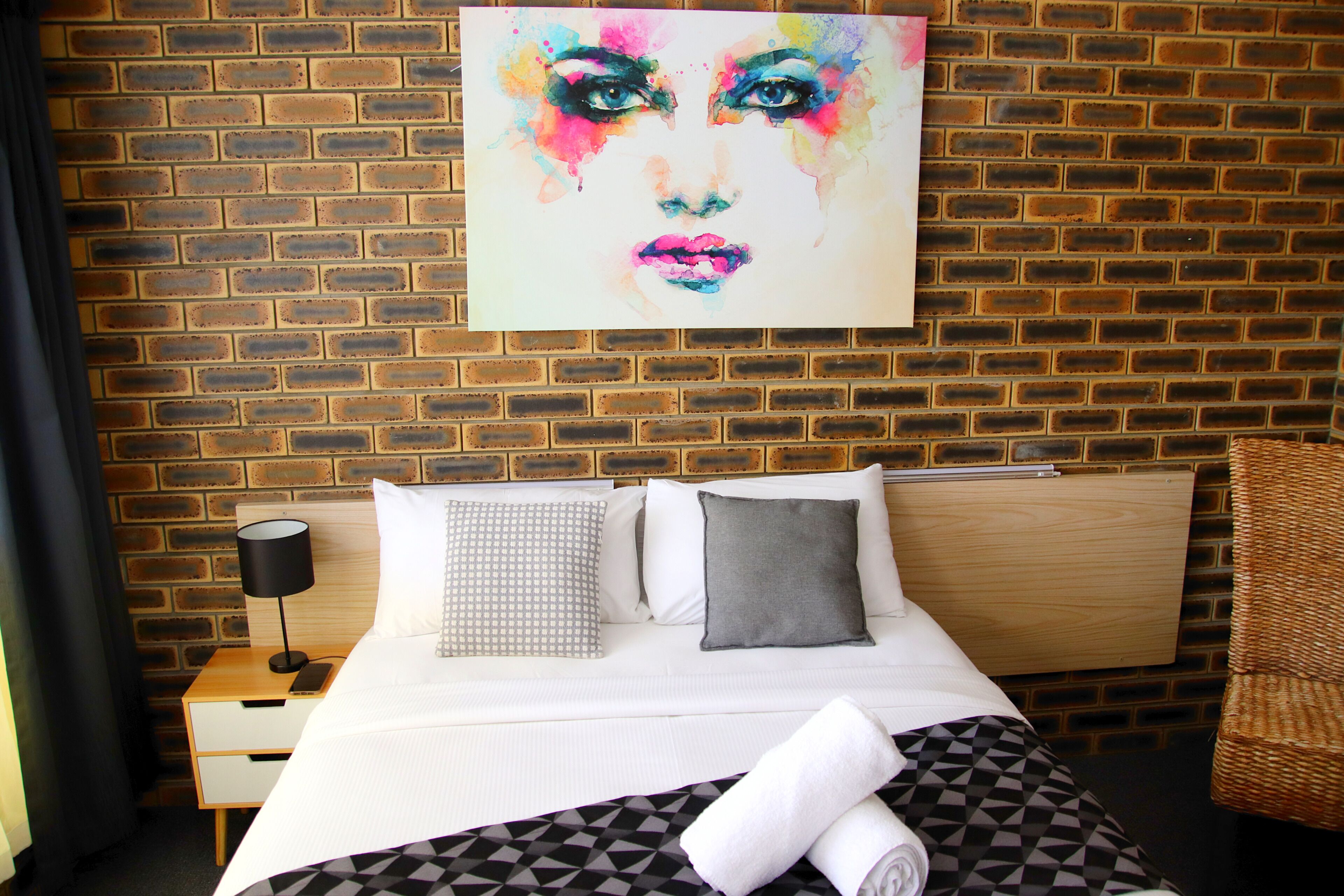 A hotel room with a brick wall, a bed with white bedding, a black and white patterned pillow, a grey pillow, a wooden nightstand with a lamp, and a wicker chair.