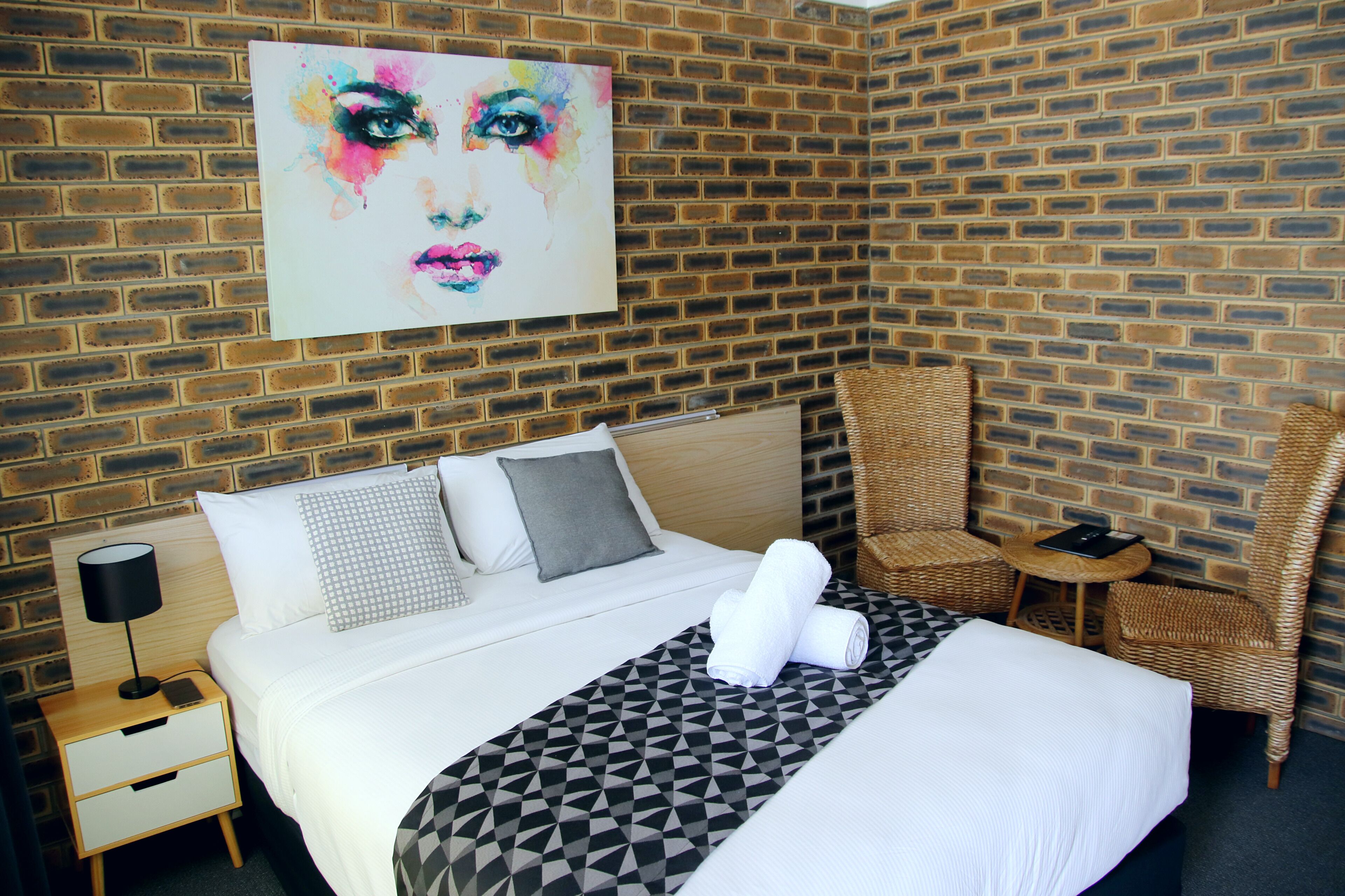 A hotel room with a brick wall, a bed with a geometric-patterned bedspread, two pillows, a nightstand with a lamp, and a wicker chair.
