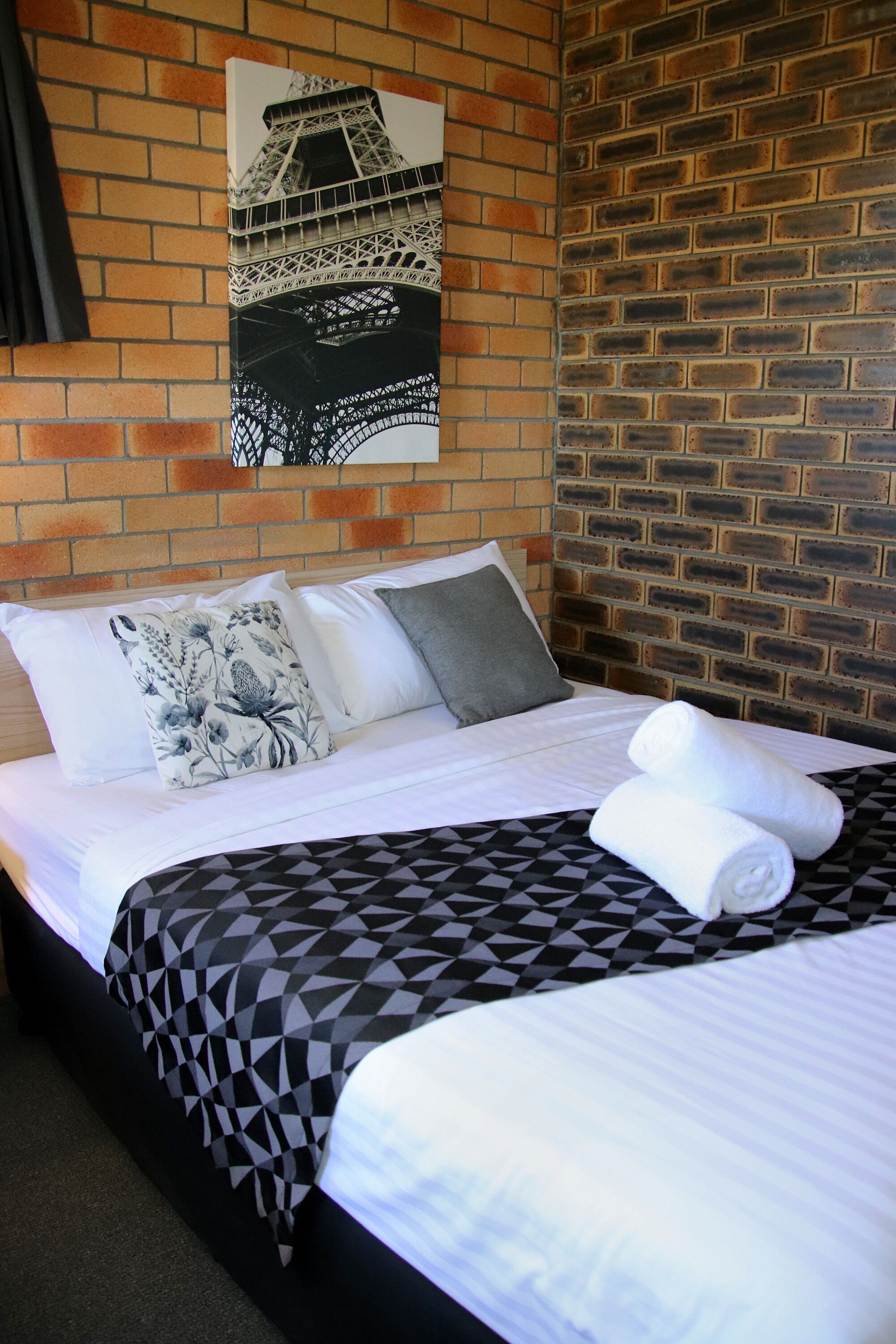 Ein Hotelzimmer mit Backsteinwand, einem Bett mit geometrischem Muster auf der Tagesdecke, zwei Kissen und einem gefalteten Handtuch.