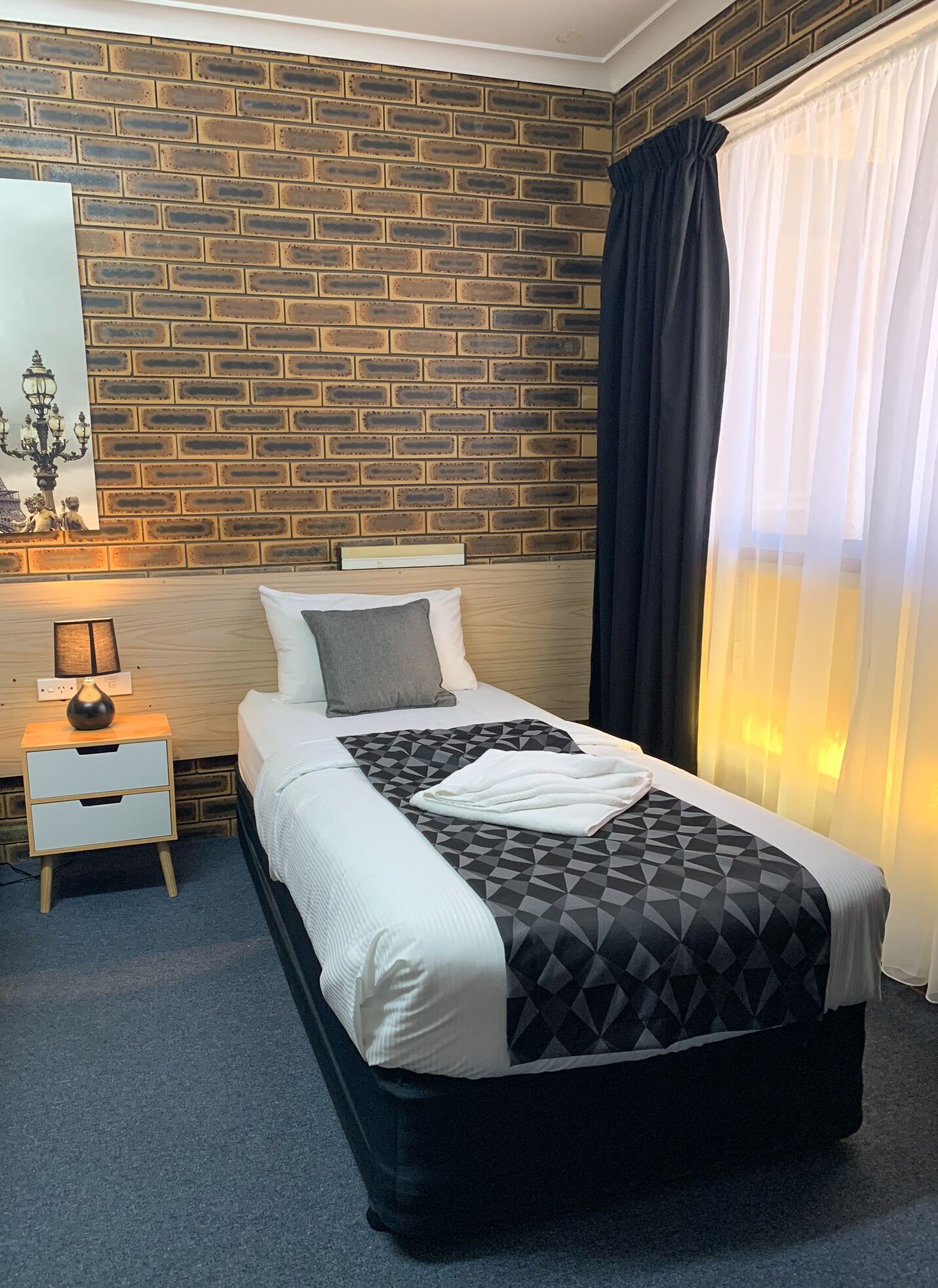 Een hotelkamer met een metselwerkmuur, een bed met een beddegoed met geometrische patronen, een nachtkastje met een lamp en een raam met dunne gordijnen.