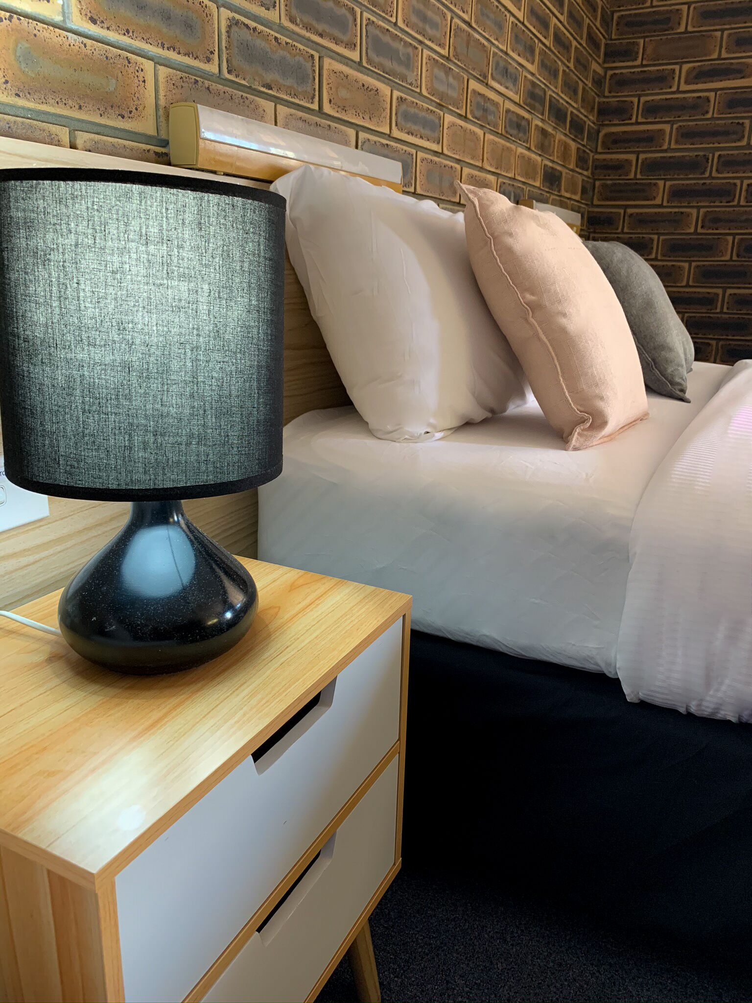 A neatly made bed with pillows, a bedside table with a lamp, and a brick-patterned wall.