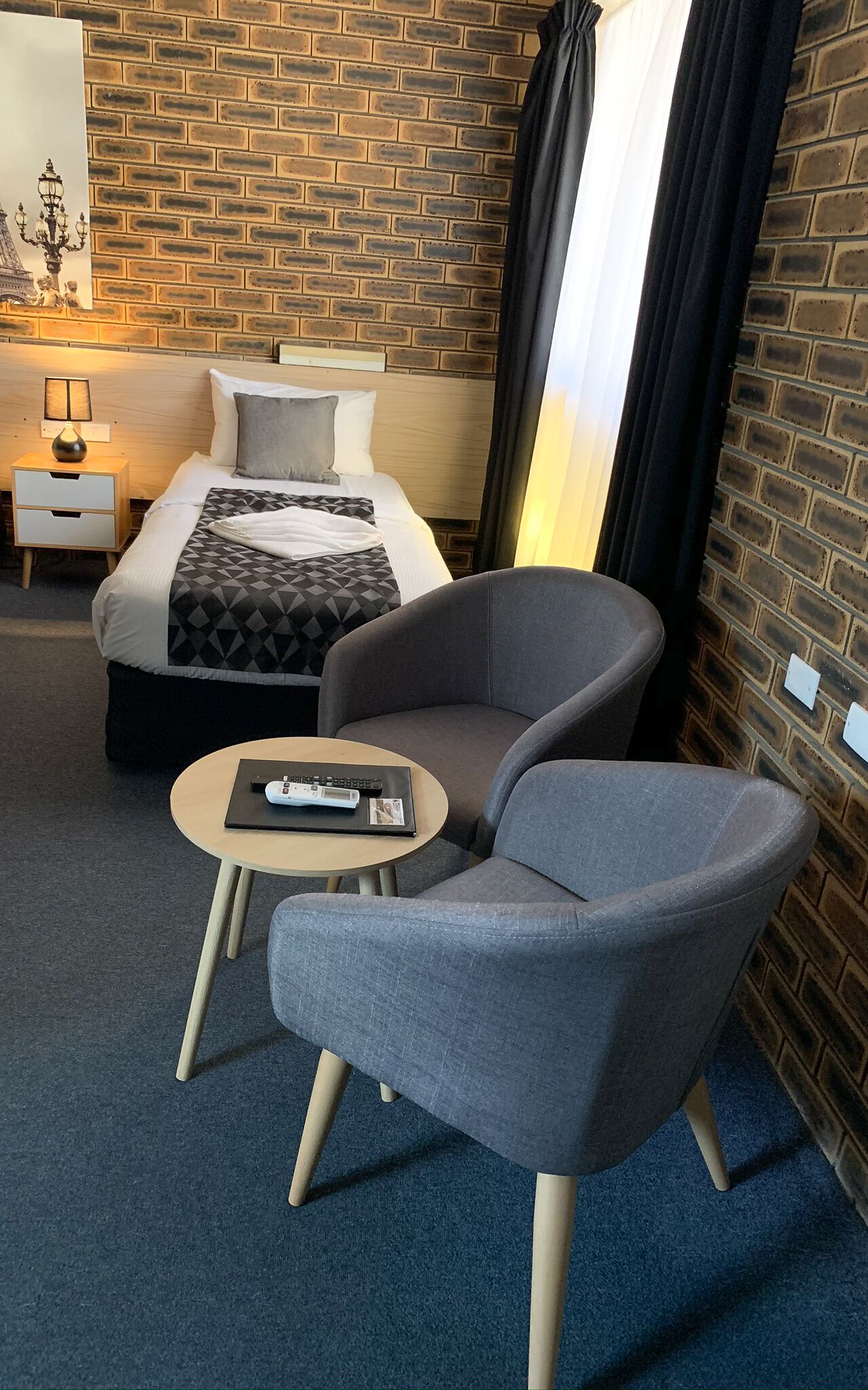 Una habitación de hotel con una pared de ladrillo, una cama con un edredón de estampado geométrico, un sillón gris, una mesita redonda con un control remoto y una mesita de noche con una lámpara.