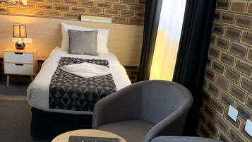 Una habitación de hotel con una pared de ladrillo, una cama con un edredón de estampado geométrico, un sillón gris, una mesita redonda con un control remoto y una mesita de noche con una lámpara.