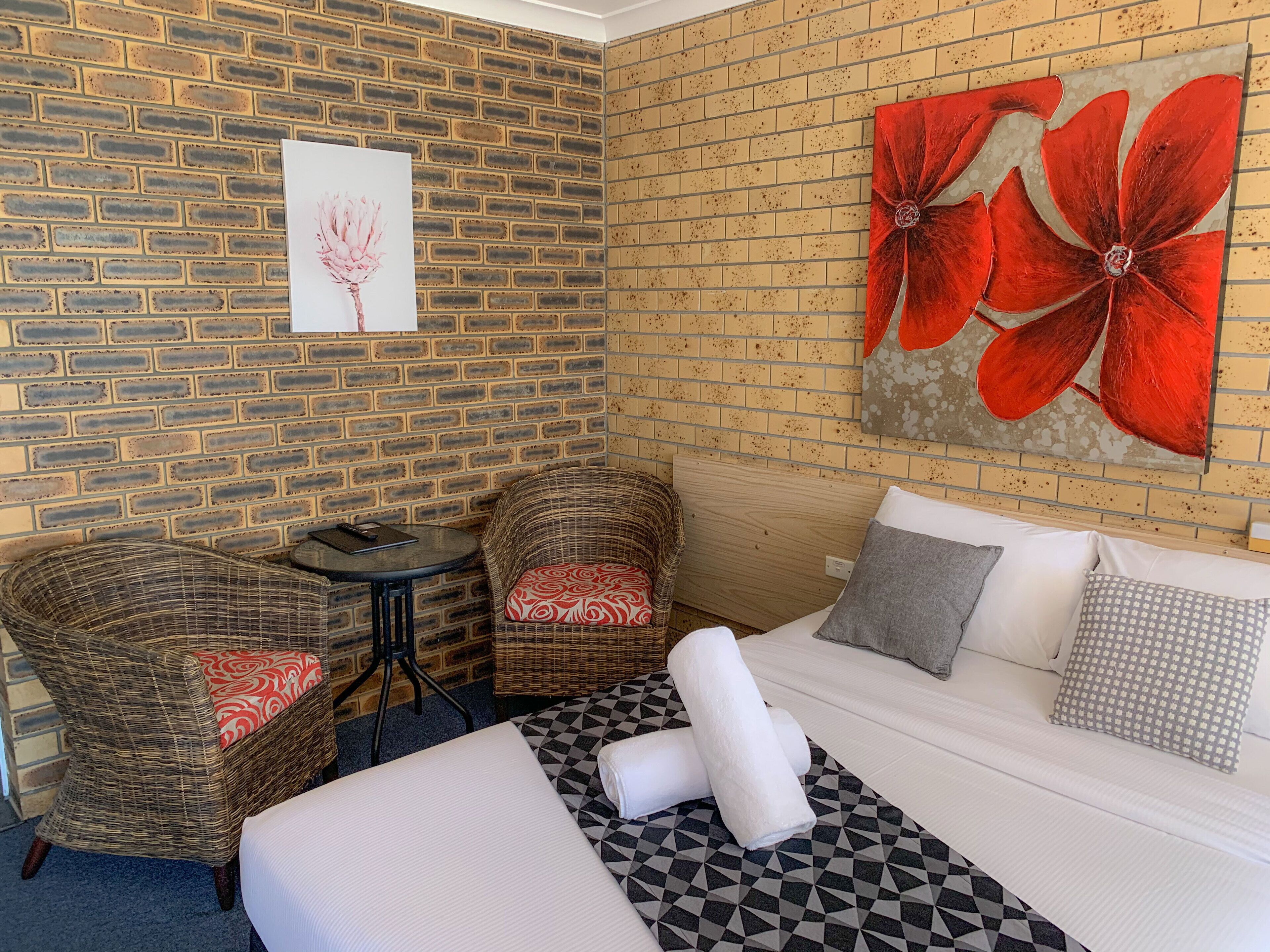 A hotel room with a brick wall, a large bed with patterned pillows, two wicker chairs, and a small round table.