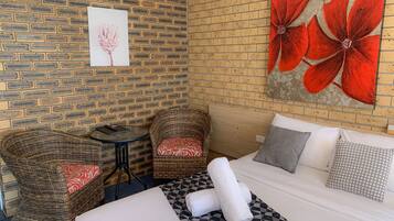 A hotel room with a brick wall, a large bed with patterned pillows, two wicker chairs, and a small round table.