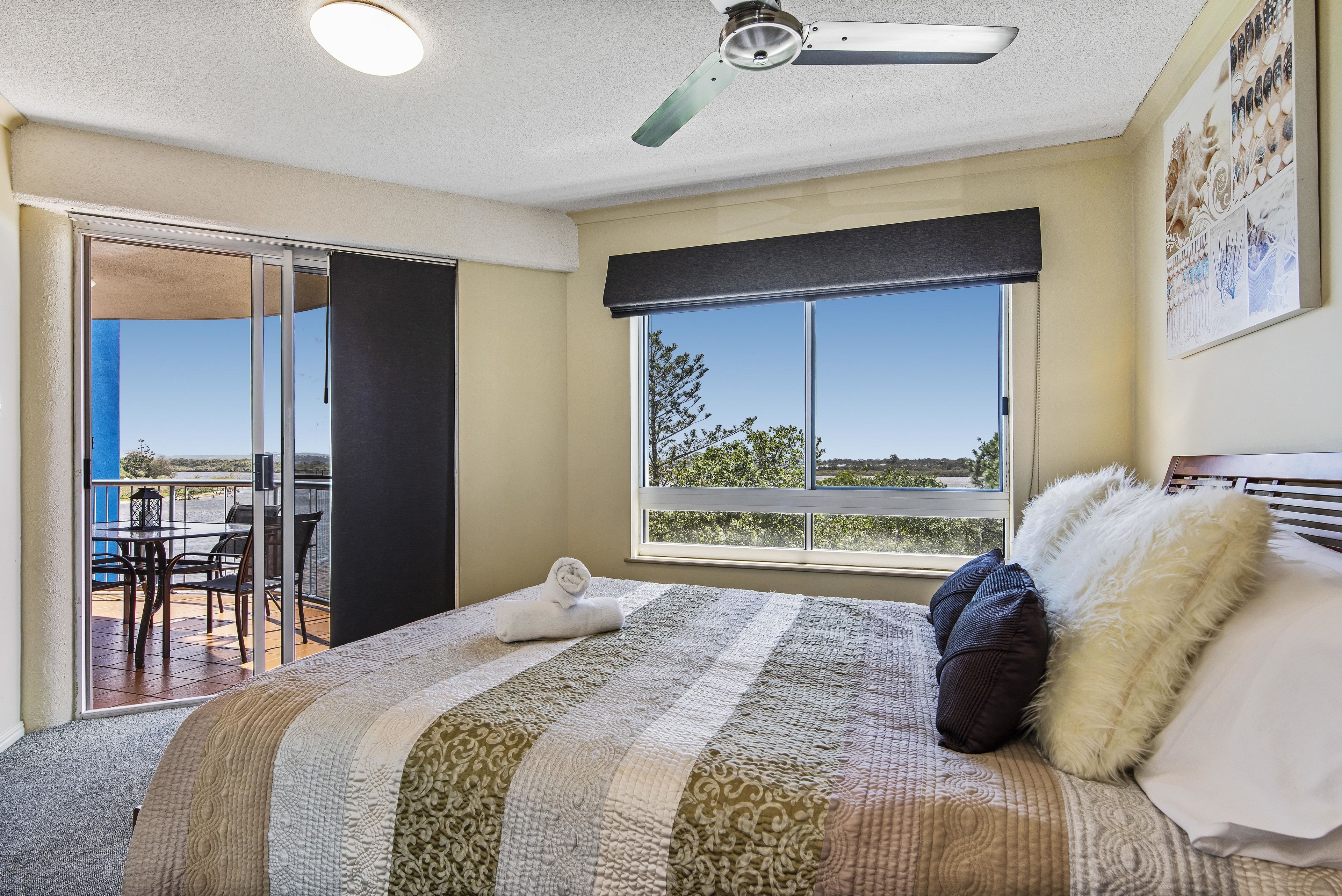 Un dormitorio con una cama grande, ventilador de techo, ventana con vistas y balcón con mobiliario exterior.