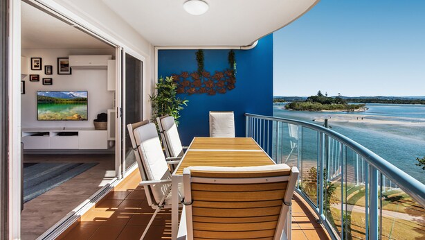 A balcony with a wooden table and chairs, a TV in the background, and a view of a body of water. - Reflections Holiday Apartments (Maroochydore)