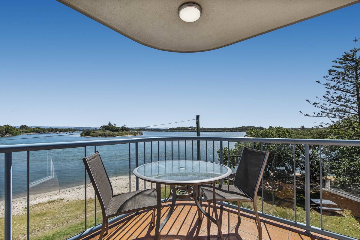 A balcony with a glass table and chairs, overlooking a body of water and a distant island.