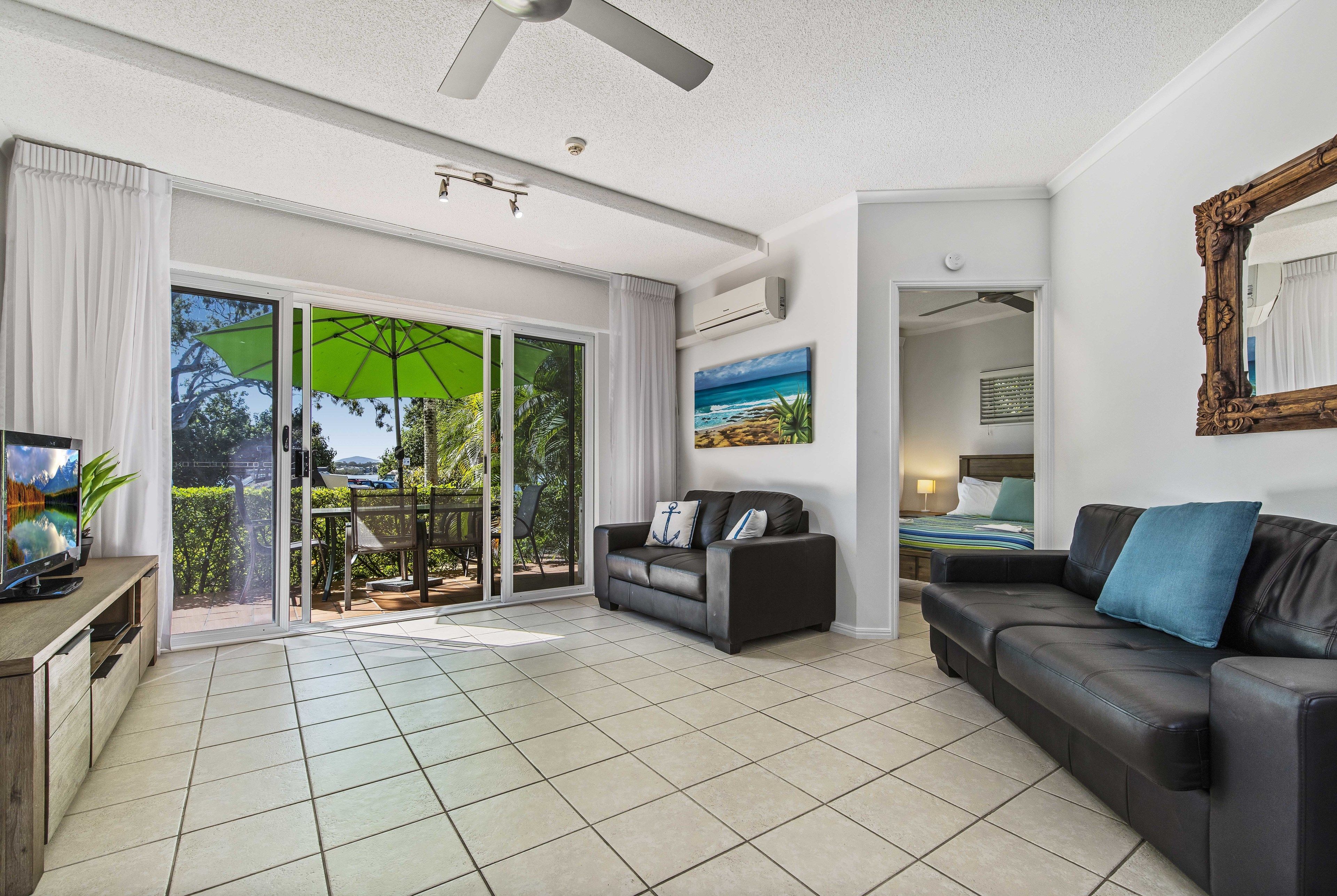 A spacious living room with a large sliding glass door leading to a balcony, a black leather sofa, a television, and a ceiling fan.