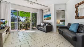 A spacious living room with a large sliding glass door leading to a balcony, a black leather sofa, a television, and a ceiling fan.