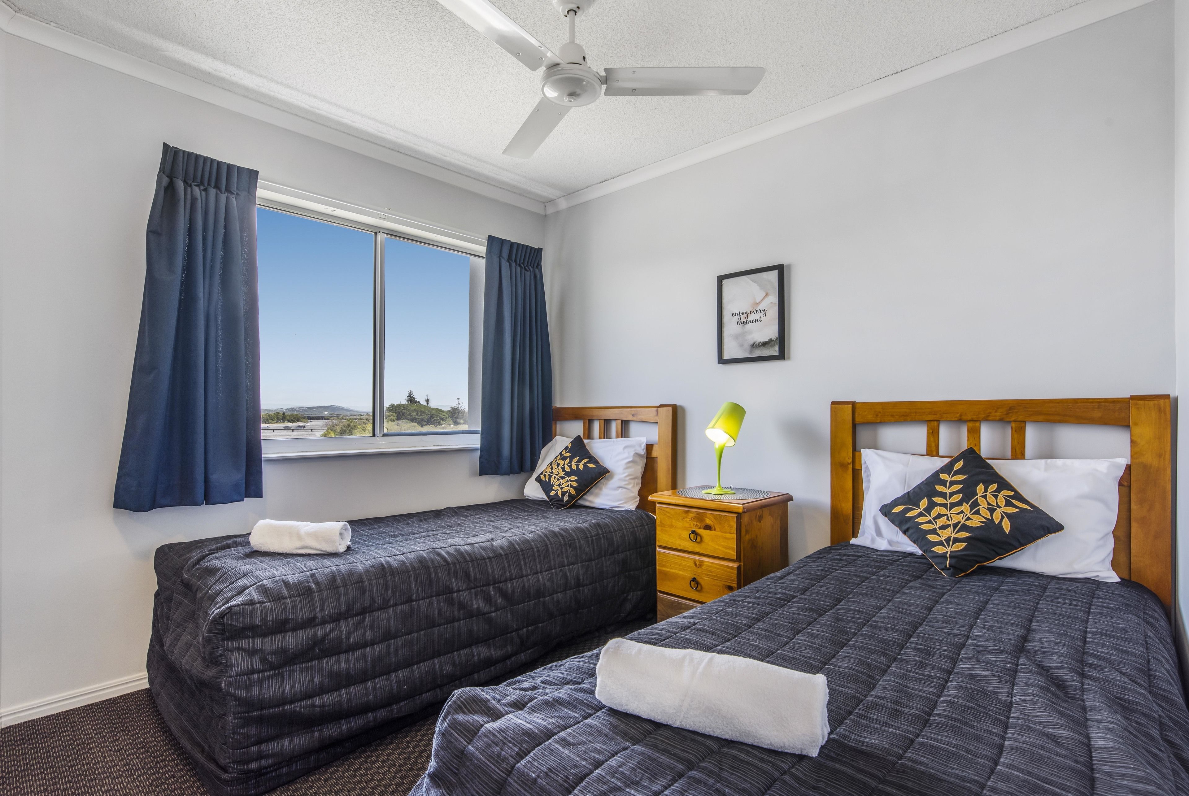 A hotel room with two beds, a window with curtains, a ceiling fan, a bedside table with a lamp, and a framed picture on the wall.