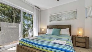 A bedroom with a large window, a bed with a striped bedspread, a wooden headboard, a nightstand with a lamp, and a view of palm trees. - Reflections Holiday Apartments (Maroochydore)