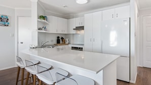 A modern kitchen with white cabinetry, a large island with bar stools, and a built-in oven. - Reflections Holiday Apartments (Maroochydore)