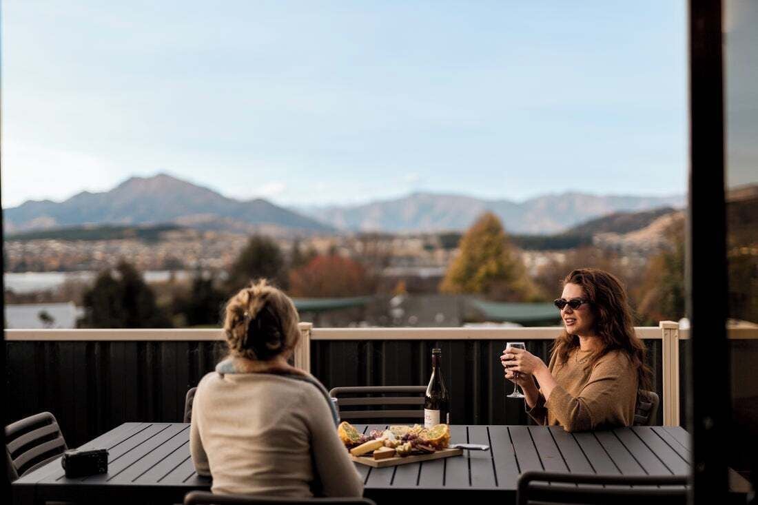 Two people seated at a table with a bottle of wine and a plate of food, overlooking a mountainous landscape.