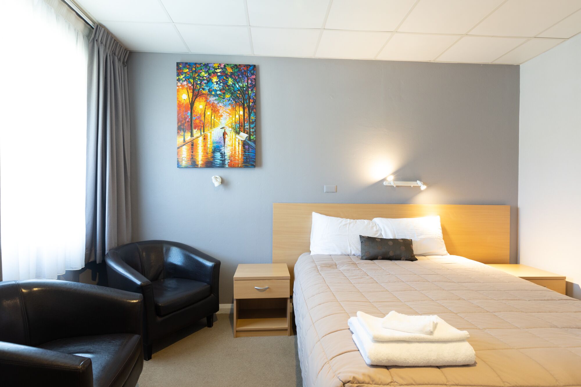 A hotel room with a bed, a chair, a nightstand, a painting on the wall, and a window with curtains.