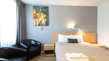 A hotel room with a bed, a chair, a nightstand, a painting on the wall, and a window with curtains.
