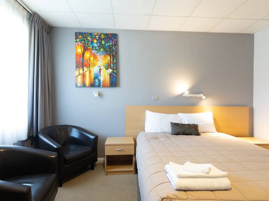 A hotel room with a bed, a chair, a nightstand, a painting on the wall, and a window with curtains.