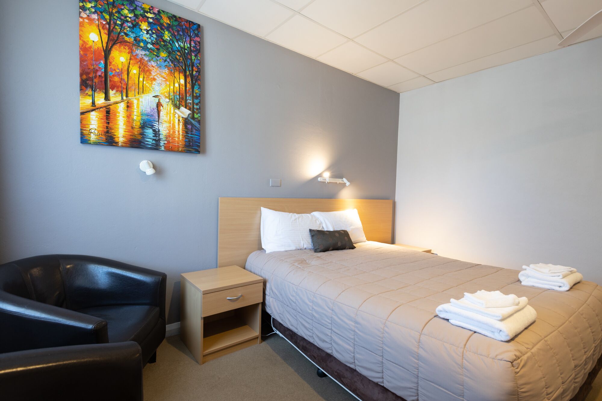 Ett hotellrum med en säng, ett nattduksbord, en stol, en tavla och en vägglampa.