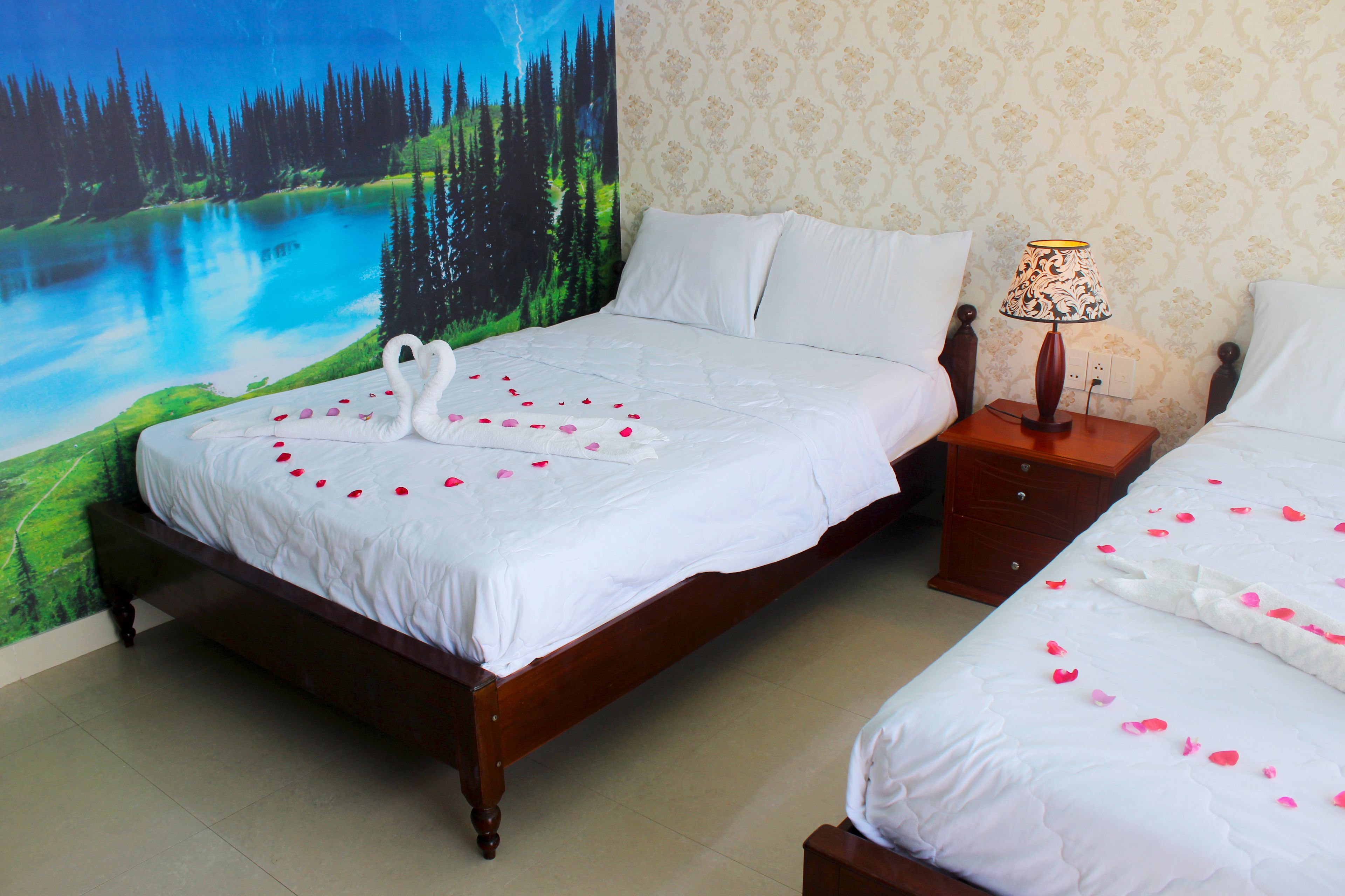 A double bed with white linens and rose petals, a nightstand with a lamp, and a forest mural on the wall.