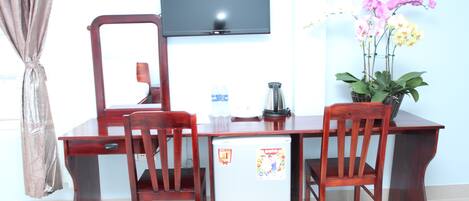 A wooden dining table with chairs, a small refrigerator, a television mounted on the wall, and a potted orchid plant.