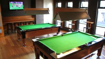 A pool table in a room with wooden flooring and a television displaying a sports game.