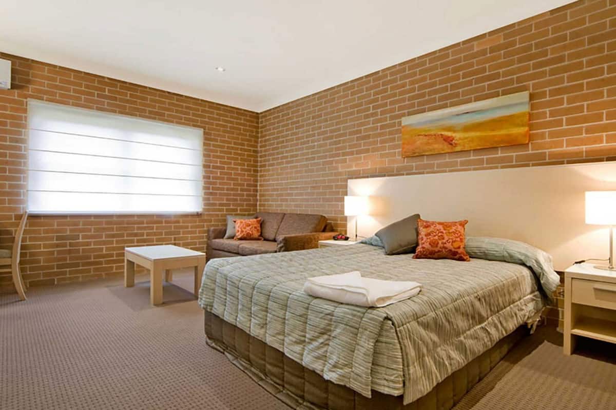 A bedroom with a brick wall, a bed with a quilt, a nightstand, a lamp, a window with blinds, and a painting.