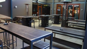 An outdoor patio area with multiple wooden tables and chairs, a central seating area with a high table, and a glass door entrance. - The Imperial Motel (Bowral)