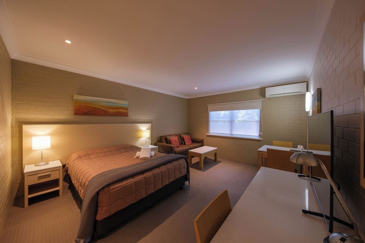 A hotel room with a bed, bedside tables, a desk, a chair, a TV, and a window with blinds.