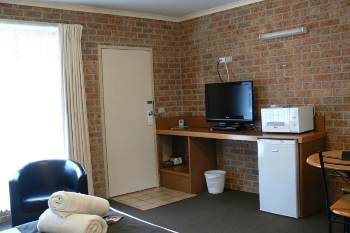 A room with a brick wall, a television, a microwave, a small refrigerator, a desk, a chair, and a door.