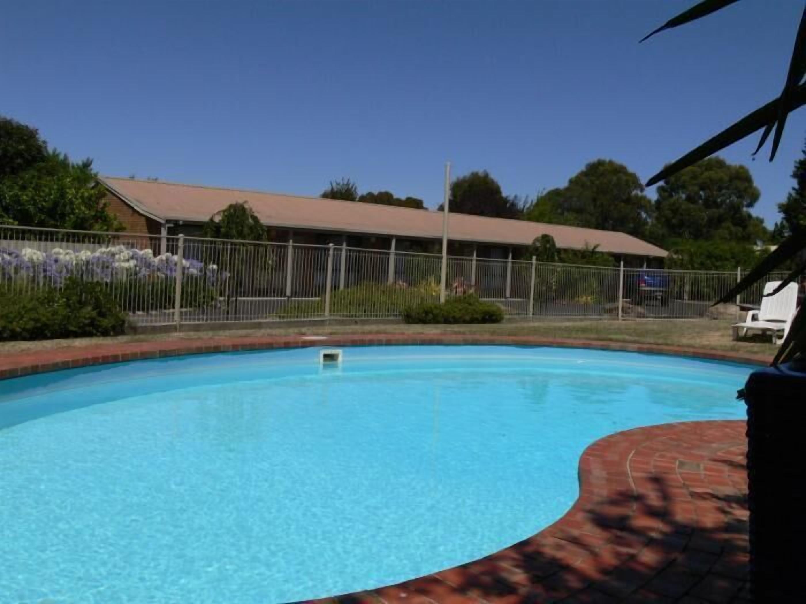 A clear blue swimming pool with a brick edge, surrounded by a fence and greenery.