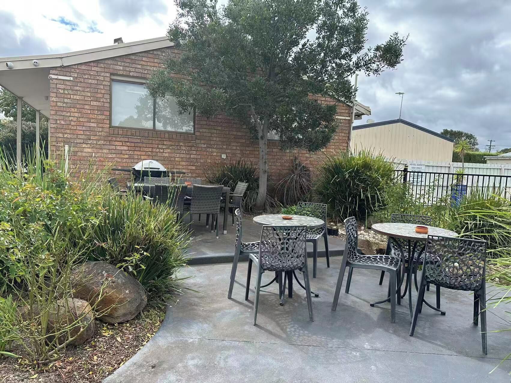 Une terrasse extérieure avec des tables et des chaises rondes, entourée de plantes et bordée d’un bâtiment en briques.