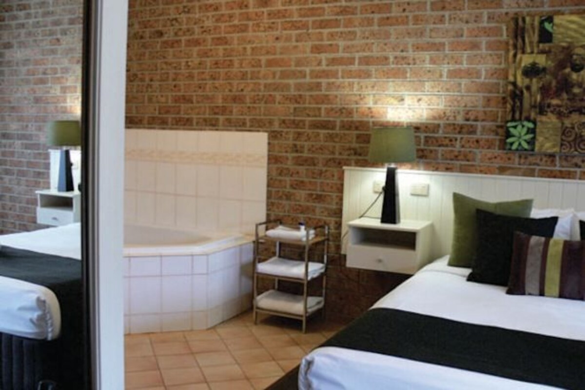 A hotel room with a brick wall, a white bathtub, a bed with multiple pillows, and a nightstand with a lamp.