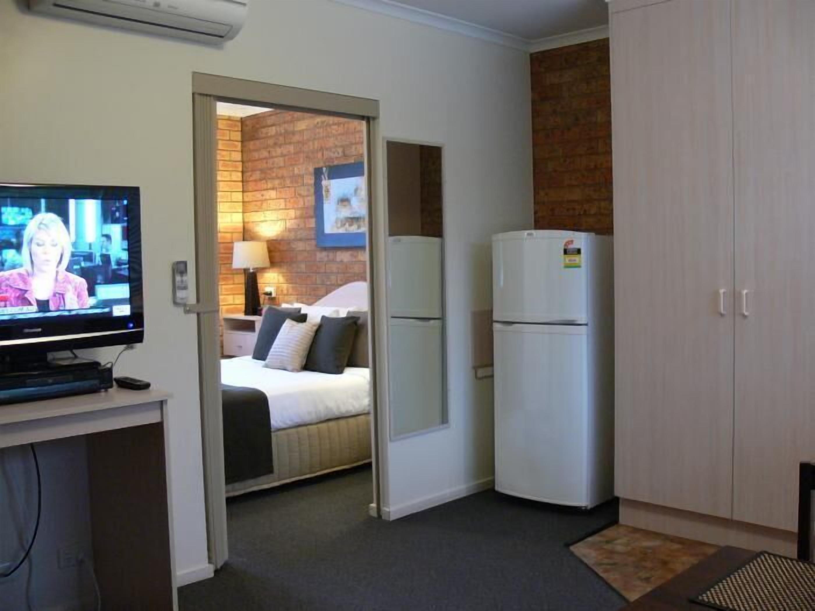 Quarto de hotel com televisão, cama, frigorífico e parede de tijolos.