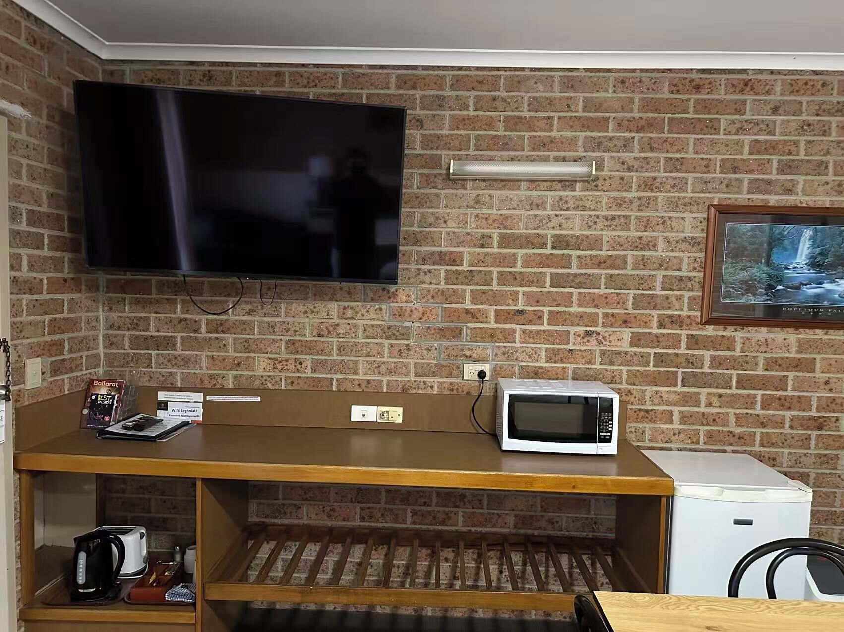 Habitación con pared de ladrillo, televisor de pantalla plana sobre un escritorio de madera, microondas blanco y refrigerador pequeño.