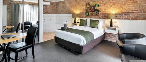 Una habitación de hotel con pared de ladrillo, cama con colcha verde, escritorio con silla, una mesita y un sofá.