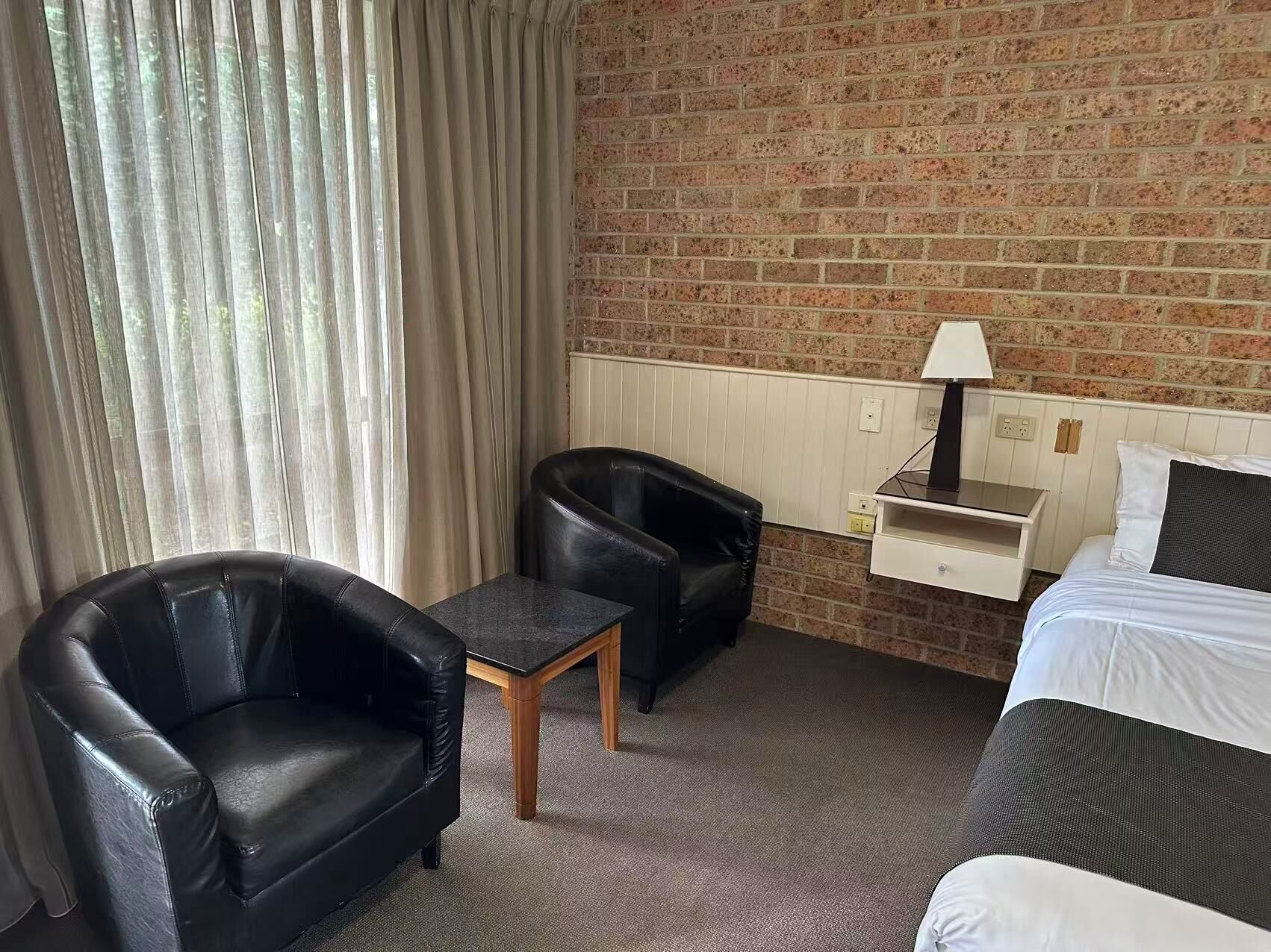 A hotel room with a brick wall, a bed, two black leather chairs, a small wooden table, and a nightstand with a lamp.