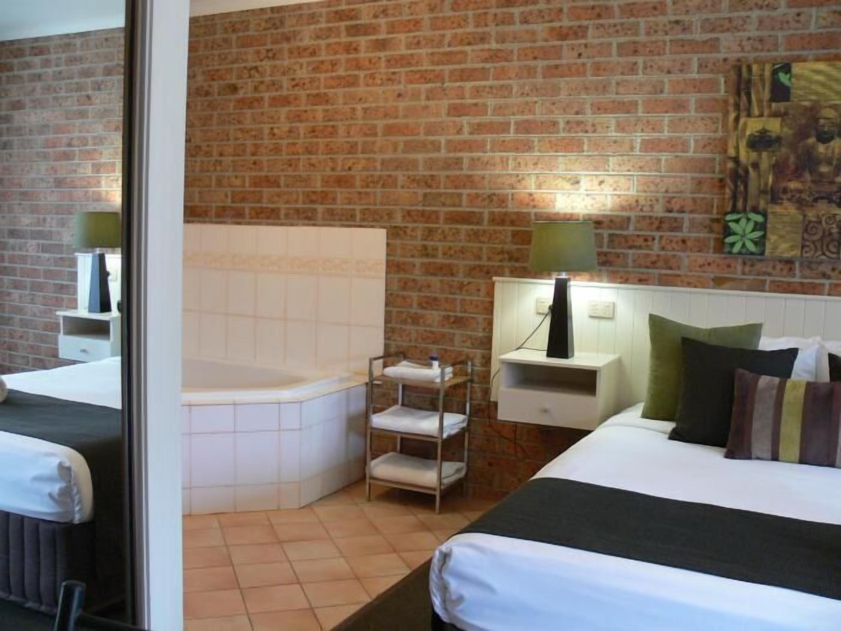 A hotel room with a brick wall, a large window, a bed, a nightstand, a lamp, and a small shelf.