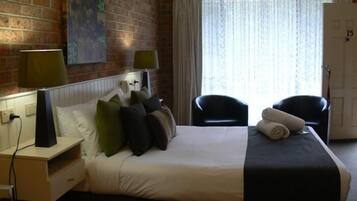 A hotel room with a brick wall, a large bed with multiple pillows, two chairs, a nightstand with a lamp, and a window with curtains.