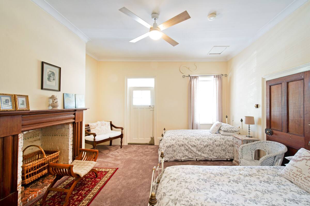 A bedroom with two beds, a fireplace, a ceiling fan, and a window with curtains.