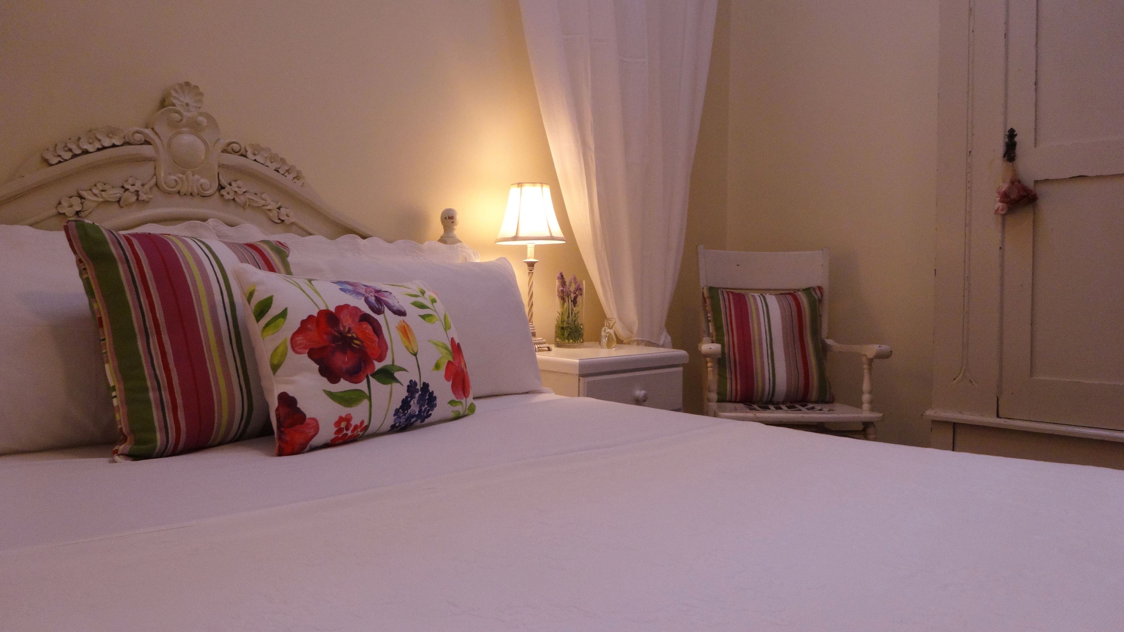 Una cama bien hecha con almohadas florales, una lámpara de noche y una silla con cojines a rayas.