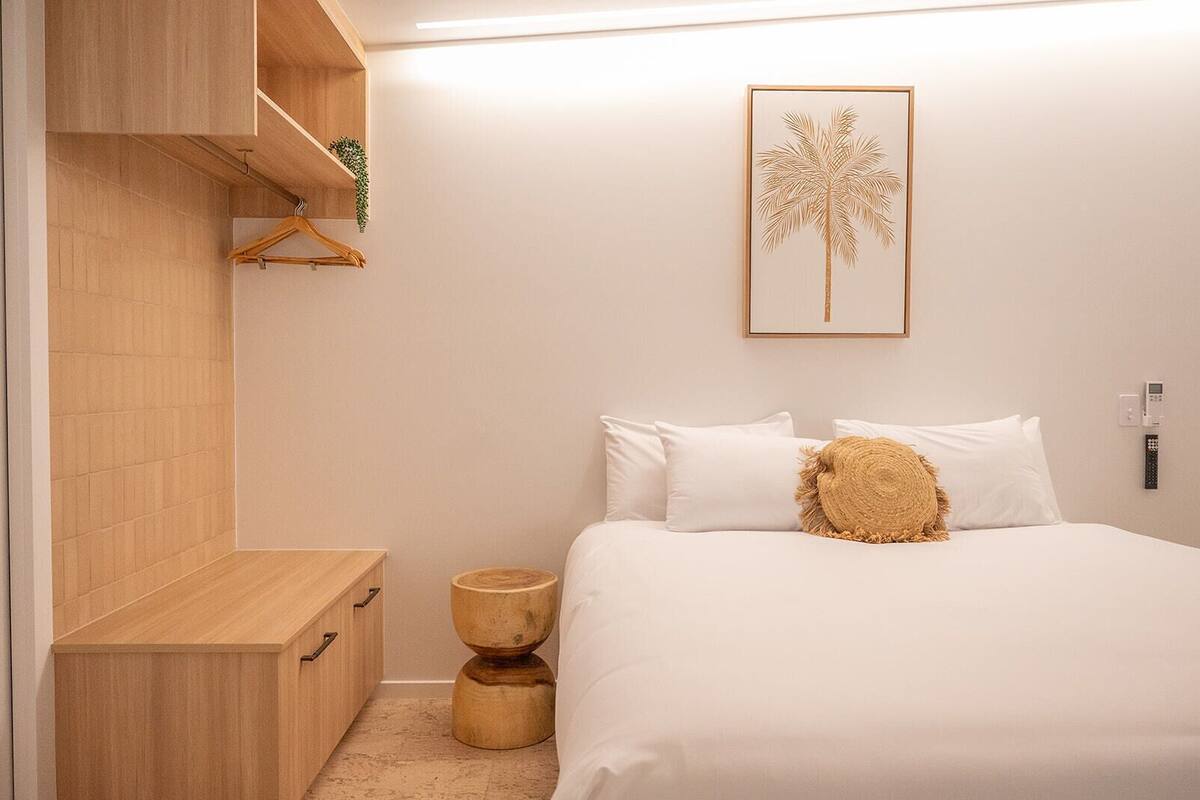 A bedroom with a bed, white pillows, a wooden bedside table, a wall-mounted shelf, a small round wooden stool, and a framed picture of a palm tree.