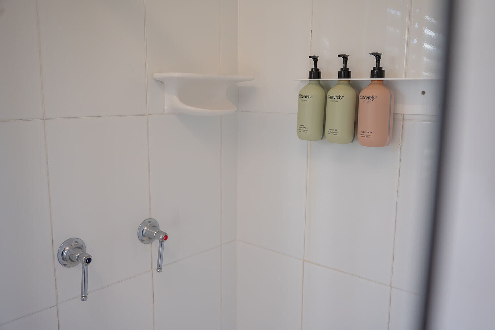A bathroom with white tiles, a shower control panel, and three bottles of Sincerely shampoo and conditioner on a shelf.