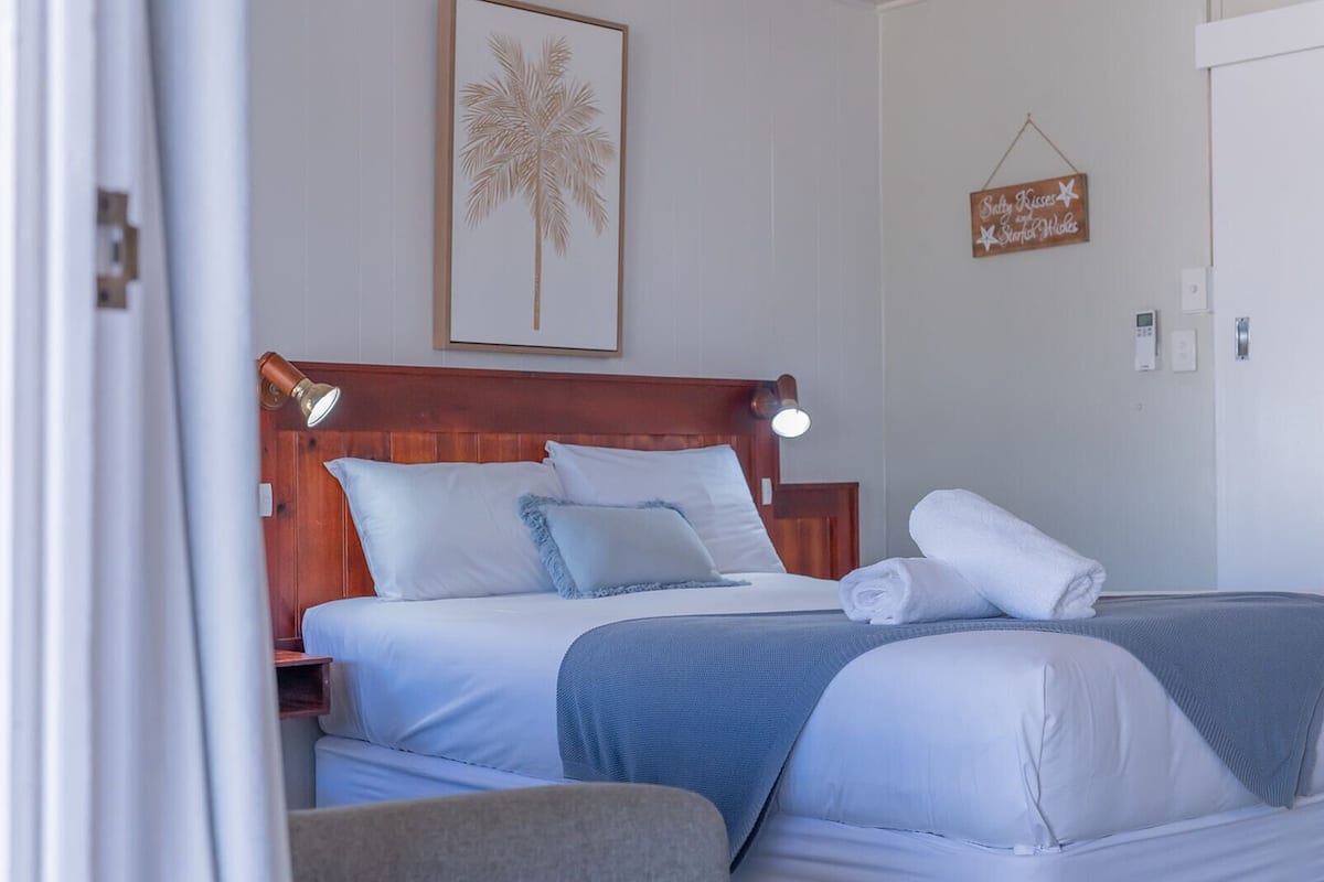 A hotel room with a wooden headboard, a bed with white and blue bedding, a rolled towel on the bed, and a wall-mounted sign.