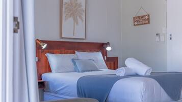 A hotel room with a wooden headboard, a bed with white and blue bedding, a rolled towel on the bed, and a wall-mounted sign.
