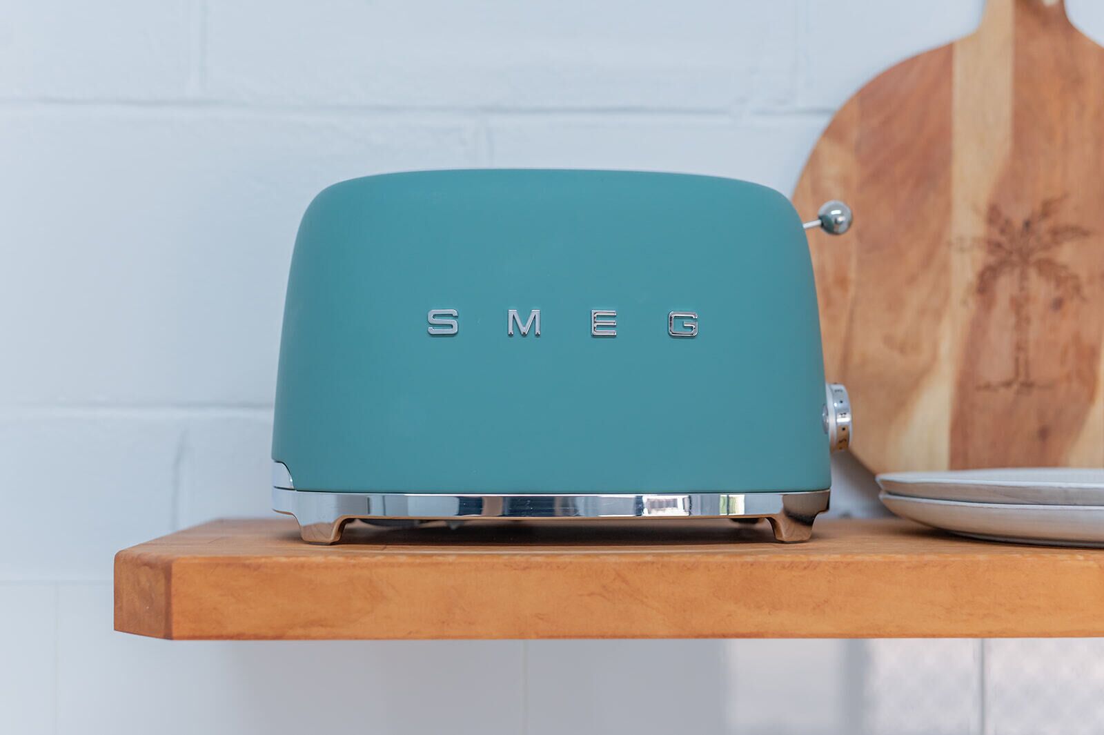 A teal-colored toaster with the brand 'SMEG' on a wooden shelf.
