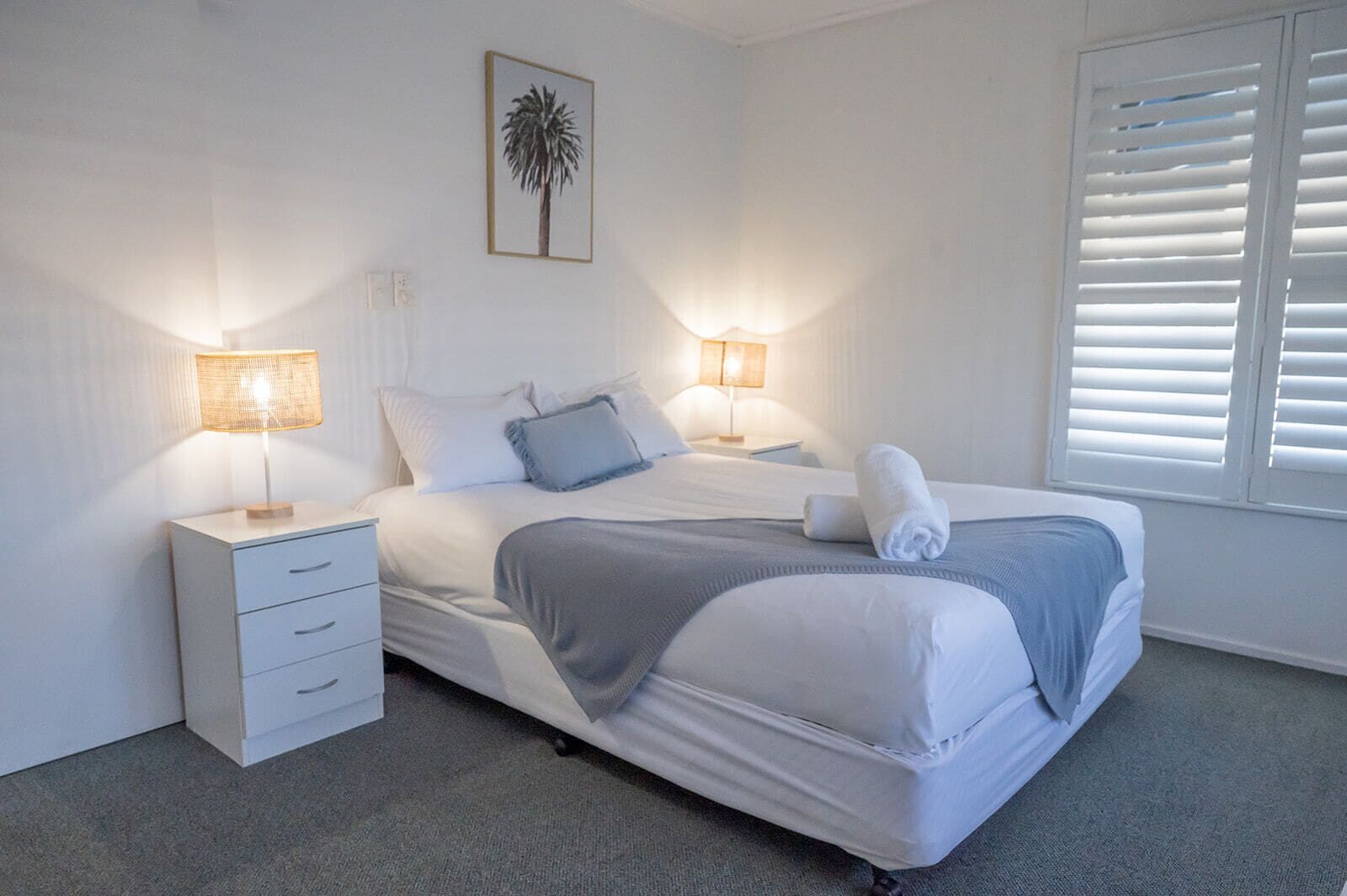 Un dormitorio ordenado con cama, dos mesitas de noche, lámpara, un cuadro en la pared y ventana con persianas.