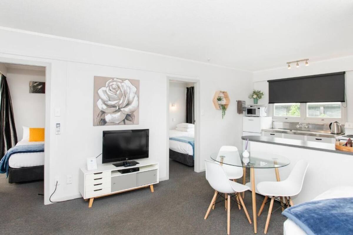 A modern hotel room with a TV, a glass dining table, white chairs, a large flower painting, and a kitchen area.